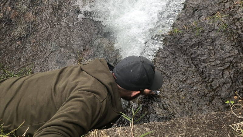 Graduate student Matthew Verosloff, hangs by one arm off a dirt ledge as he leans his body over the edge of a river.